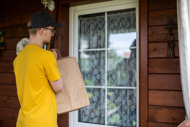 Joven repartidor en camiseta amarilla llevando bolsas de papel delante de la puerta de casa - Foto, Imagen