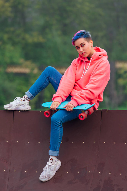 Portrait of a trendy pretty young girl with short colored hair and nose piercing dressed in pink hoodie and jeans sitting next to the skateboard coart with her blue plastic skateboard. - Foto, imagen