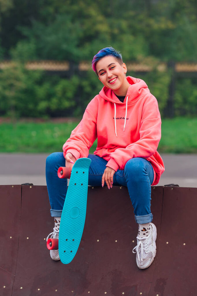 Portrait of a trendy pretty young girl with short colored hair and nose piercing dressed in pink hoodie and jeans sitting next to the skateboard coart with her blue plastic skateboard. - Foto, Imagen