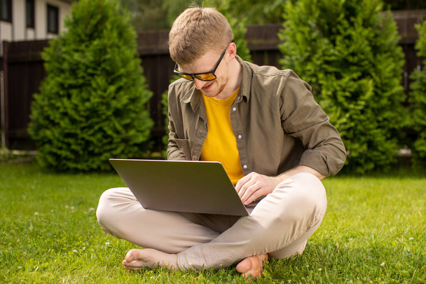 Młody szczęśliwy człowiek odpoczywający na trawie w parku z laptopem w rękach - Zdjęcie, obraz