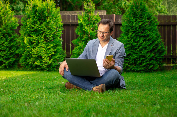 Upset middle aged businessman sits outdoors with laptop and mobile phone - Photo, Image