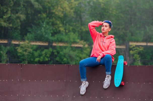 Portrait of a trendy pretty young girl with short colored hair and nose piercing dressed in pink hoodie and jeans sitting next to the skateboard coart with her blue plastic skateboard. Copy space - Foto, Imagem