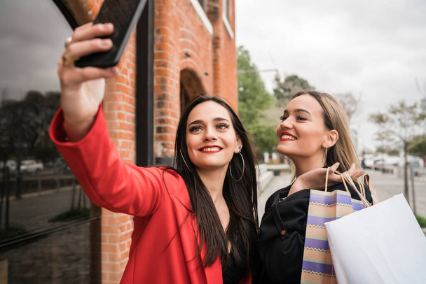 Portrait of two young friends enjoying doing shopping together while taking a selfie with phone at the street. Friendship and shopping concept. - Foto, immagini