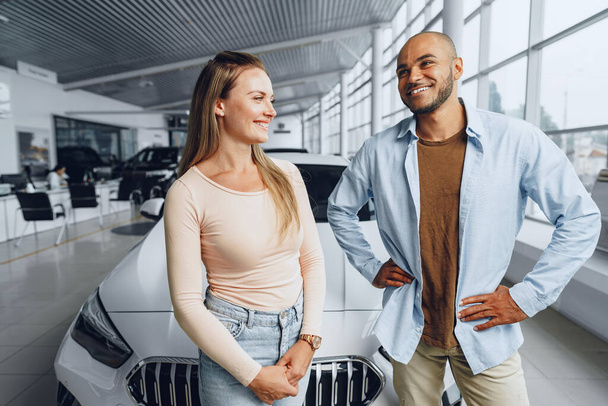 Happy couple of caucasian woman and african american man standing near their new car inside car salon - Foto, afbeelding