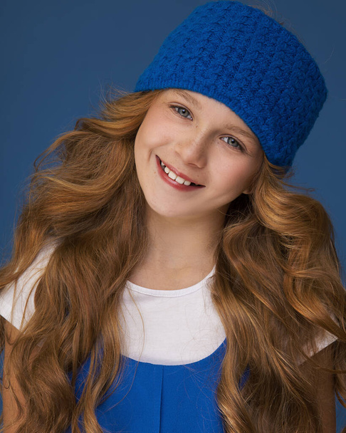 Gros plan Portrait d'une jeune adolescente de 10 ans aux cheveux roux luxueux en boucles et taches de rousseur. Souriez et regardez la caméra dans le Studio à l'arrière-plan bleu. Concept d'accessoire chapeau chaud. - Photo, image
