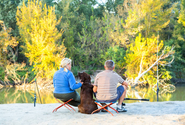 retired couple with labrador dog fishing on autumn river, sitting with dog on the folding chairs near fishing rods - Photo, Image