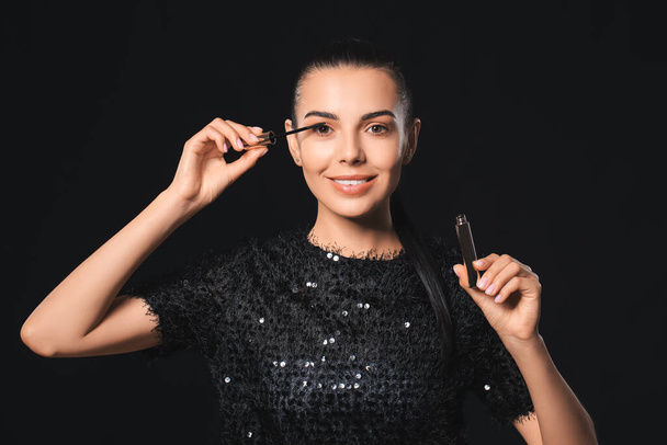 Beautiful young woman applying mascara against dark background - Foto, Imagen