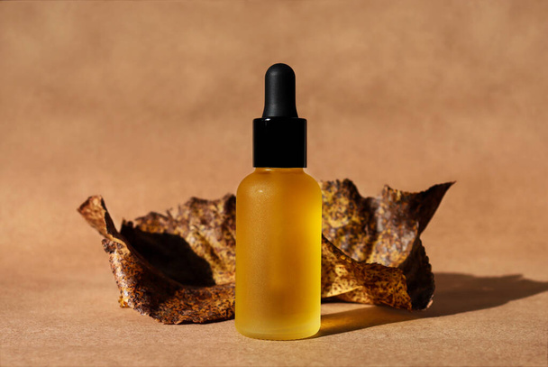 Dropper oil bottle with black cap, craft paper background, dry leaves. Horizontal monochrome brown mockup. Autumn poster. Natural organic cosmetic, lotion, essence. Biophilia, terracotta brick color - Φωτογραφία, εικόνα