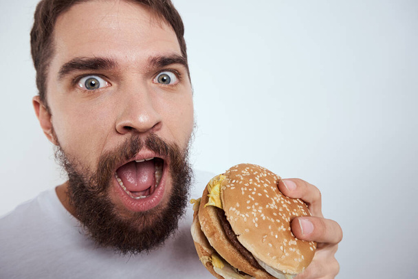 Un homme mange un hamburger sur un fond clair dans un T-shirt blanc vue rapprochée faim fast food - Photo, image