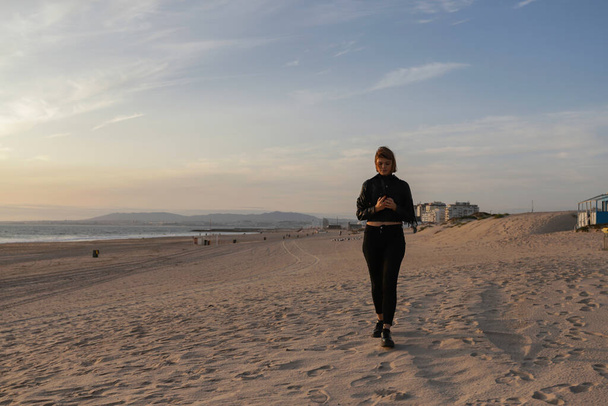Красивая кавказская женщина, идущая вдоль пляжа на Коста да Капарика недалеко от центра Лиссабона. Молодая женщина в кожаной куртке отдыхает на пляже на закате в Капарике. Португалия, 2020 год - Фото, изображение