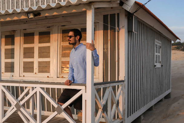 Um jovem bonito posando sob o alpendre de uma típica casa de campo ao longo da praia na Costa da Caparica, usando óculos de sol ao pôr do sol e olhando para o Oceano Atlântico. Lisboa, Portugal 2020 - Foto, Imagem