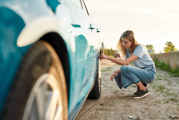 Attractive young woman checking air pressure of car tire on local road side while traveling, Girl having troubles with her auto, checking wheel after car breakdown - Valokuva, kuva