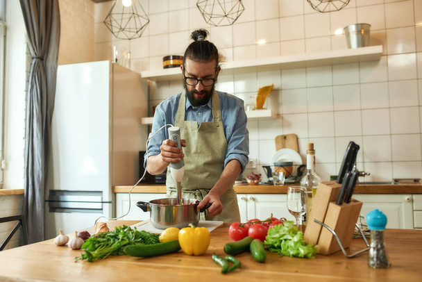 Joven, cocinero usando licuadora de manos mientras prepara comida italiana en la cocina - Foto, imagen
