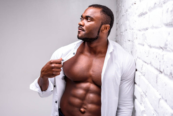 Strong african athlete in a white shirt. Posing showing perfect muscle shape near a white brick wall - Foto, Bild