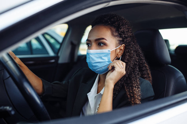 Young business woman fixing and adjusting her medical mask while sitiing in the car behind the steering wheel. Business trips during pandemic, new normal and coronavirus travel safety concept - Fotó, kép