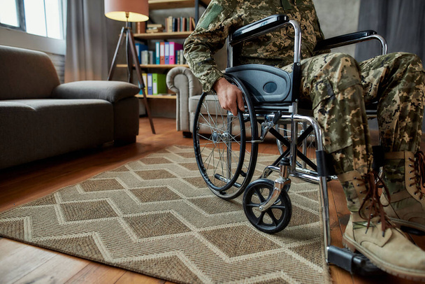 Mozgássérült. Megvágták a mozgássérült katona képét, aki tolószékben álcázta magát a terápián a nappaliban. PTSD-koncepció - Fotó, kép