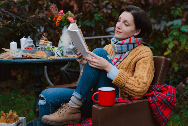 молодая женщина в коричневом пальто сидит в кресле за столом и читает книгу с клетчатой поверх головы на открытом воздухе против осенней покрасневшей листвы. - Фото, изображение