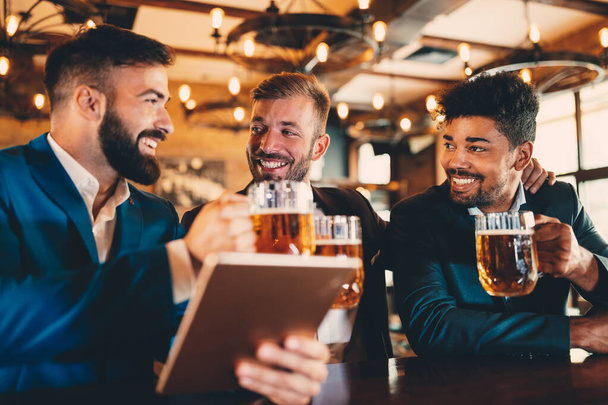 Handsome businessmen drinking beer, talking and smiling while resting at the pub - 写真・画像