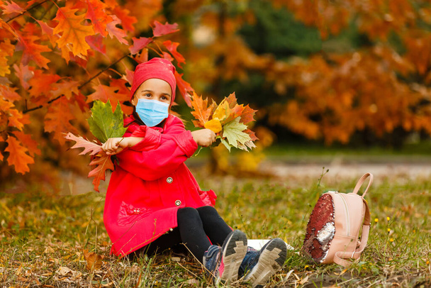 Маленькая девочка в медицинской маске предотвращает загрязнение опавших листьев в парке. - Фото, изображение