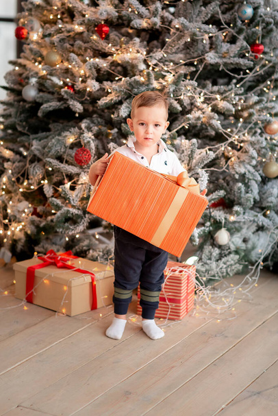 Wesoły, słodki chłopczyk z prezentami. Dzieciak trzyma duży prezent i bawi się przy drzewie rano. Wesołych Świąt i Happy Holidays koncepcja - Zdjęcie, obraz