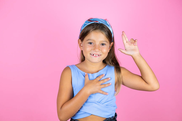 Joven niña hermosa sobre fondo rosa aislado sonriendo jurando con la mano en el pecho y los dedos hacia arriba, haciendo una promesa de lealtad juramento - Foto, Imagen