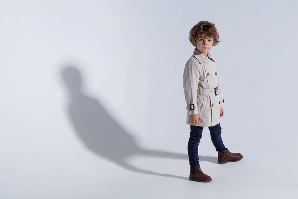 Volledige lengte van een stijlvolle schattige krullende kleine jongen, dragen in een beige loopgraaf, poseert in de studio, geïsoleerde witte achtergrond. Kopieerruimte. - Foto, afbeelding