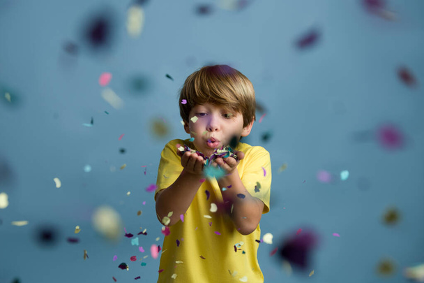 Ξανθό αγόρι διασκεδάζει φυσώντας κομφετί σε χρωματιστό μπλε φόντο. - Φωτογραφία, εικόνα