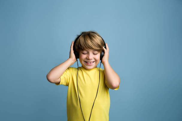 Divertido niño pequeño disfrutando de ritmos al escuchar música en los auriculares. Sonriente chico rubio en camiseta amarilla - Foto, imagen
