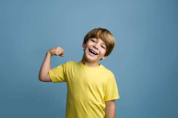 Portrait d'un garçon mignon montrant les muscles de ses bras. Garçon blond avec t-shirt jaune - Photo, image