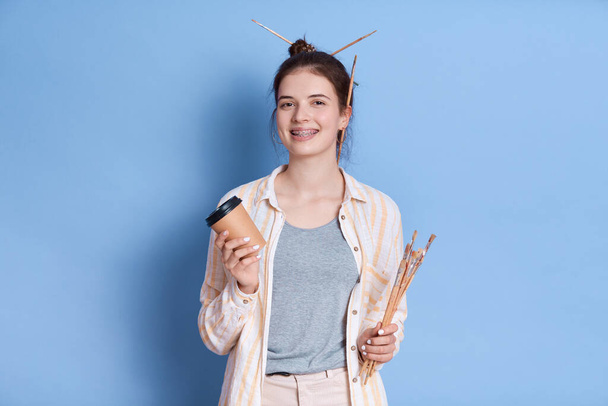 Portrait of female painter standing isolated over blue background, looking smiling directly at camera, artist wearing white shirt, holding take away coffee and brushes. - Fotografie, Obrázek