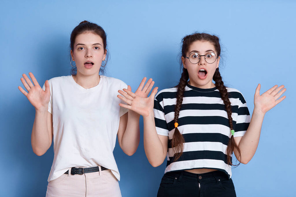 European dark haired girls posing with hands up, widely opened mouth, having shocked facial expressions, express fear, wearing casual t shirts, posing against blue wall. - Photo, Image