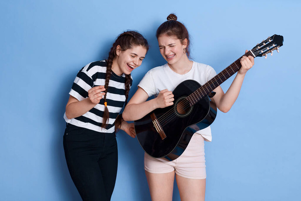 Winsome ladies with knot and pigtails singing songs and playing guitar, having fun together, preparing for music competition, posing isolated over blue background. - Foto, Bild