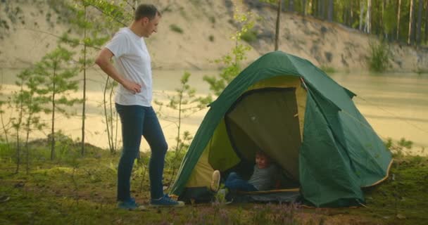 Jongen klimt in een tent aan de oever van het meer tijdens het reizen en ontspannen met zijn vader in de natuur - Video