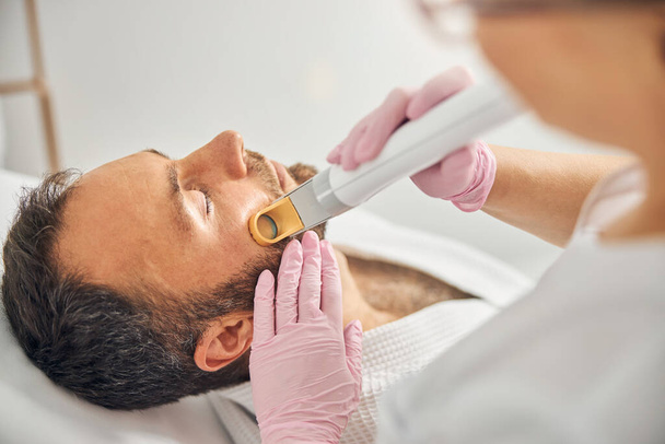 Esthéticienne enlevant les poils indésirables de la joue masculine avec dispositif laser - Photo, image