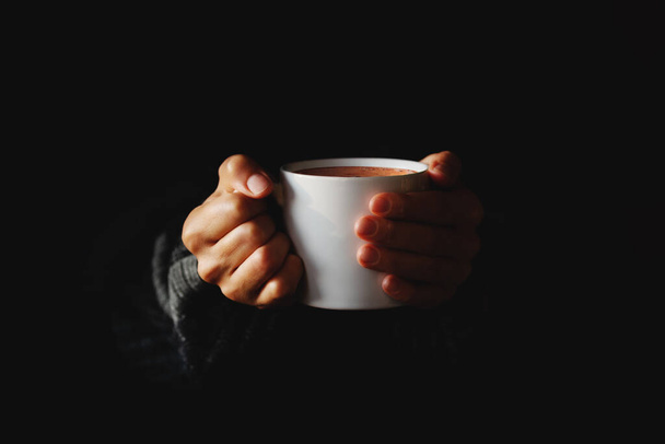 Una taza de café en las manos de las mujeres sobre el fondo negro. Mujer calentando sus manos en taza de bebida caliente en la oscuridad. Copiar espacio. Estilo mínimo. Tecla baja - Foto, Imagen