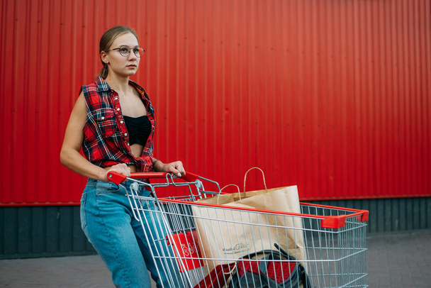 Onnellinen tyttö ostoskärryillä punaisella seinäpuodin taustalla. Nuori nainen työntää ostoskärry täynnä ostoksia ja ostoksia - Valokuva, kuva