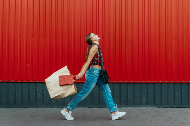 Boldog lány bevásárló papírzacskóval piros fali bolt háttér. Fiatal nő gazdaság bevásárló táska tele élelmiszerekkel és vásárlásokkal vásárlás után. - Fotó, kép