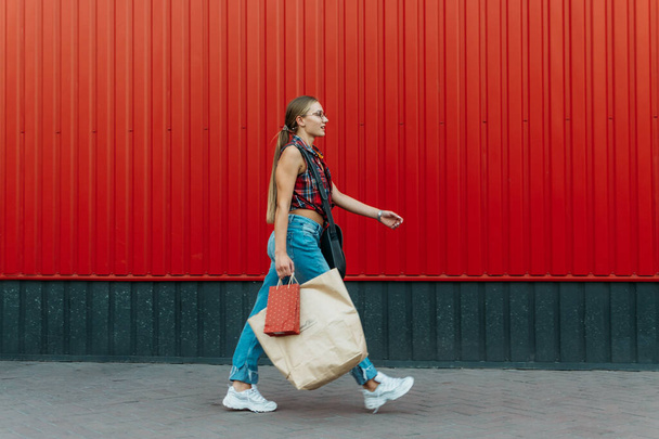 Счастливая девушка с бумажным пакетом на красном фоне магазина. Молодая женщина держит сумку с продуктами и покупками после покупок. - Фото, изображение