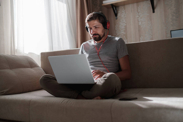 Άνδρας στον καναπέ με ακουστικά και σημειωματάριο στο σπίτι. οι νέοι hipster φορούν ακουστικά σπουδάζουν online παρακολουθώντας webinar podcast σε laptop ακούγοντας μαθήματα εκμάθησης ή μουσικής, μαθαίνοντας έννοια - Φωτογραφία, εικόνα