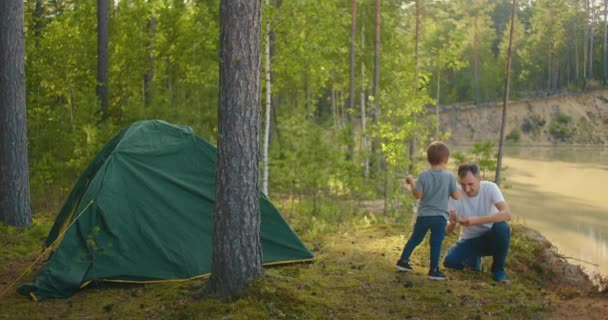 De jongen helpt zijn vader bij het opzetten en opzetten van een tent in het bos. Kinderen onderwijzen en samen reizen in een tentenkamp - Video