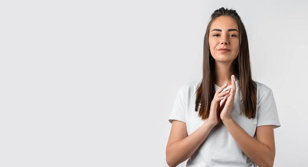 Photo of charismatic Pensive European young woman with long chestnut hair keeps hands together near chin, has cute expression, wears white t shirt. Copy space for your text - Photo, Image
