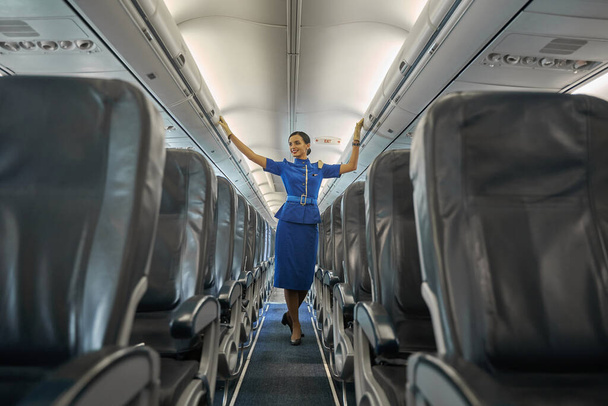 Συνοπτική επαγγελματική αεροσυνοδός χαμογελαστή στην καμπίνα του αεροσκάφους - Φωτογραφία, εικόνα