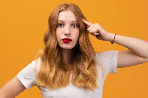スタジオは白いTシャツを着て、頭に指を指して波状の赤毛の若い女性を自信を持って撮影しました。思考の概念。賢く考えなさい. - 写真・画像