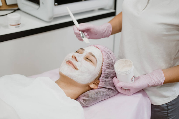プロのエステティシャンがスパの美容センターでお客様の顔に顔マスクを適用します。若いです女性取得フェイシャルケアによって美容室にきび治療、顔剥離マスク. - 写真・画像