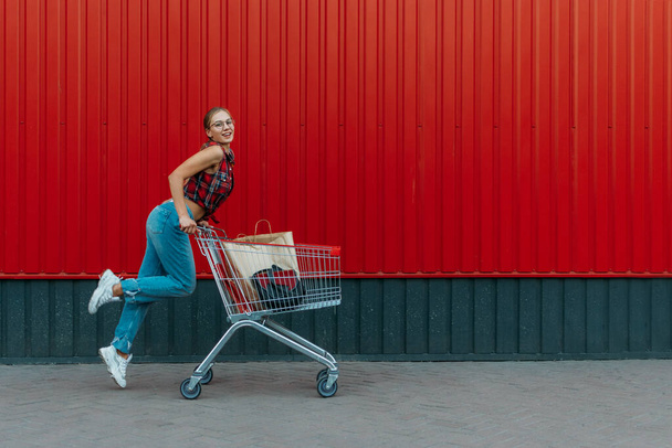 Happy girl with shopping cart on red wall shop background. Young woman pushing a shopping cart full of groceries and purchases - Photo, Image