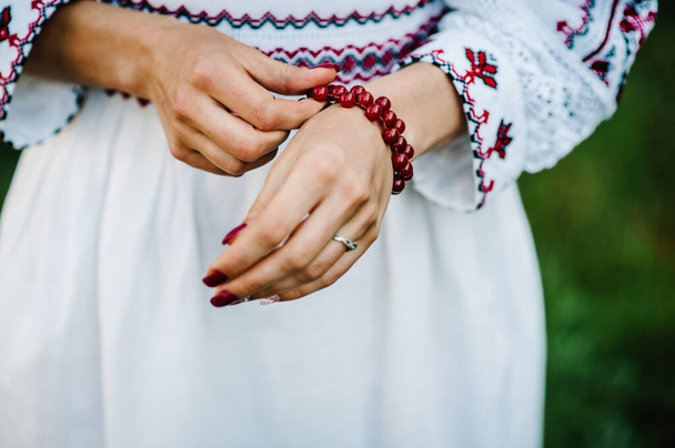 View of female hand with red lacquer on nails and a beautiful wears bracelet of gemstones on rustic style autumn. Ukrainian style: bride in embroidered clothes on nature. - Фото, изображение