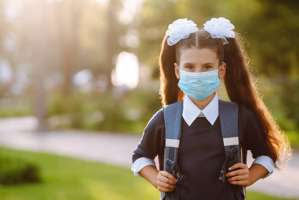 Schoolgirl in protective sterile mask when returning to school during the COVID-19 quarantine. Modern pupil with a backpack at sunset. - Foto, immagini