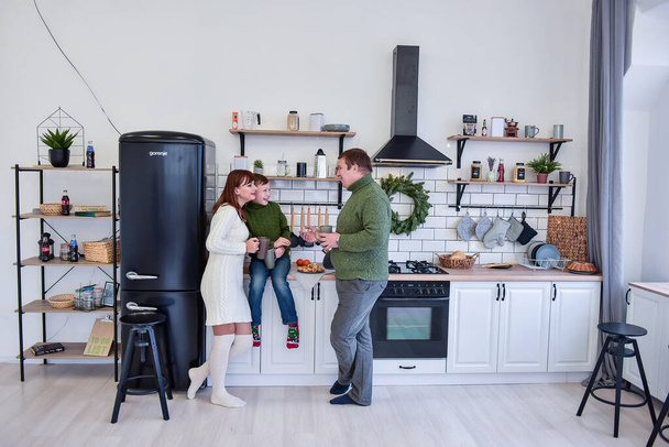 Happy family in the white kitchen in a Scandinavian style, drinking hot drinks and laughing. A young woman hugs her son, a man holds a gray cup in his hands. Minimalistic christmas wreath. Copy space  - Photo, image