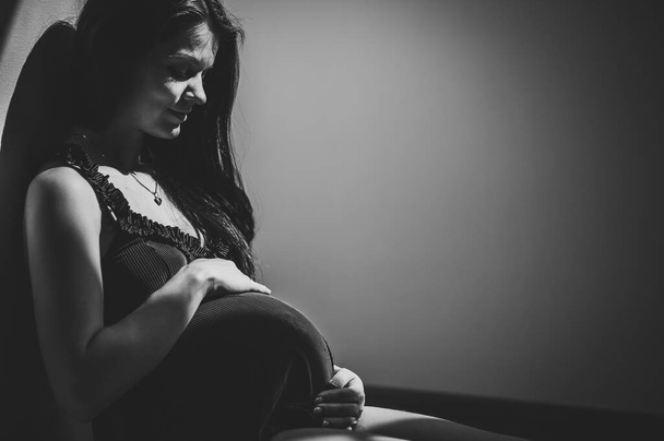 Bekliyorum bebeğim. Hamile kadın pencerenin yanında oturuyor ve elleri yuvarlak bir göbeğe ve mideye sarılıyor. Yakın plan. Dokuz ay. Bebek partisi. Annelik kavramı. Siyah beyaz fotoğraf. - Fotoğraf, Görsel