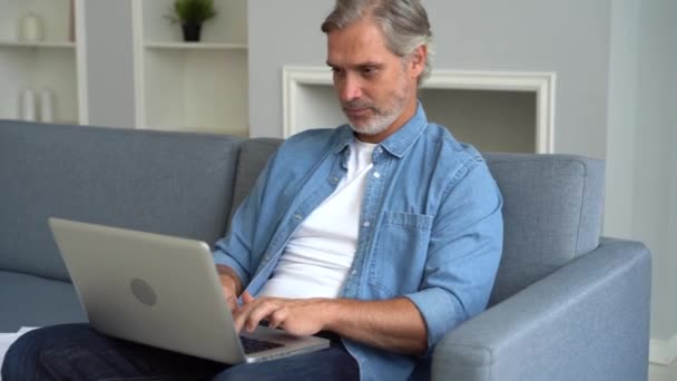 Attractive man using his laptop sitting at home on the sofa in the living room - Záběry, video